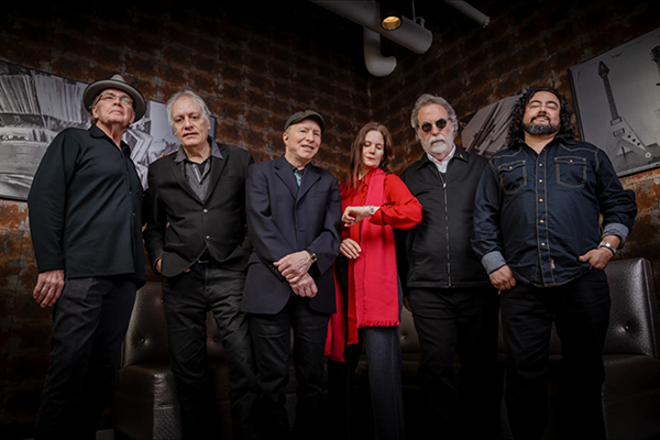 10000-maniacs → Visit Hopewell / Prince George VA Six-member band posing in a dim studio with a brick wall; the woman in a red scarf stands center, flanked by four men in dark jackets.
