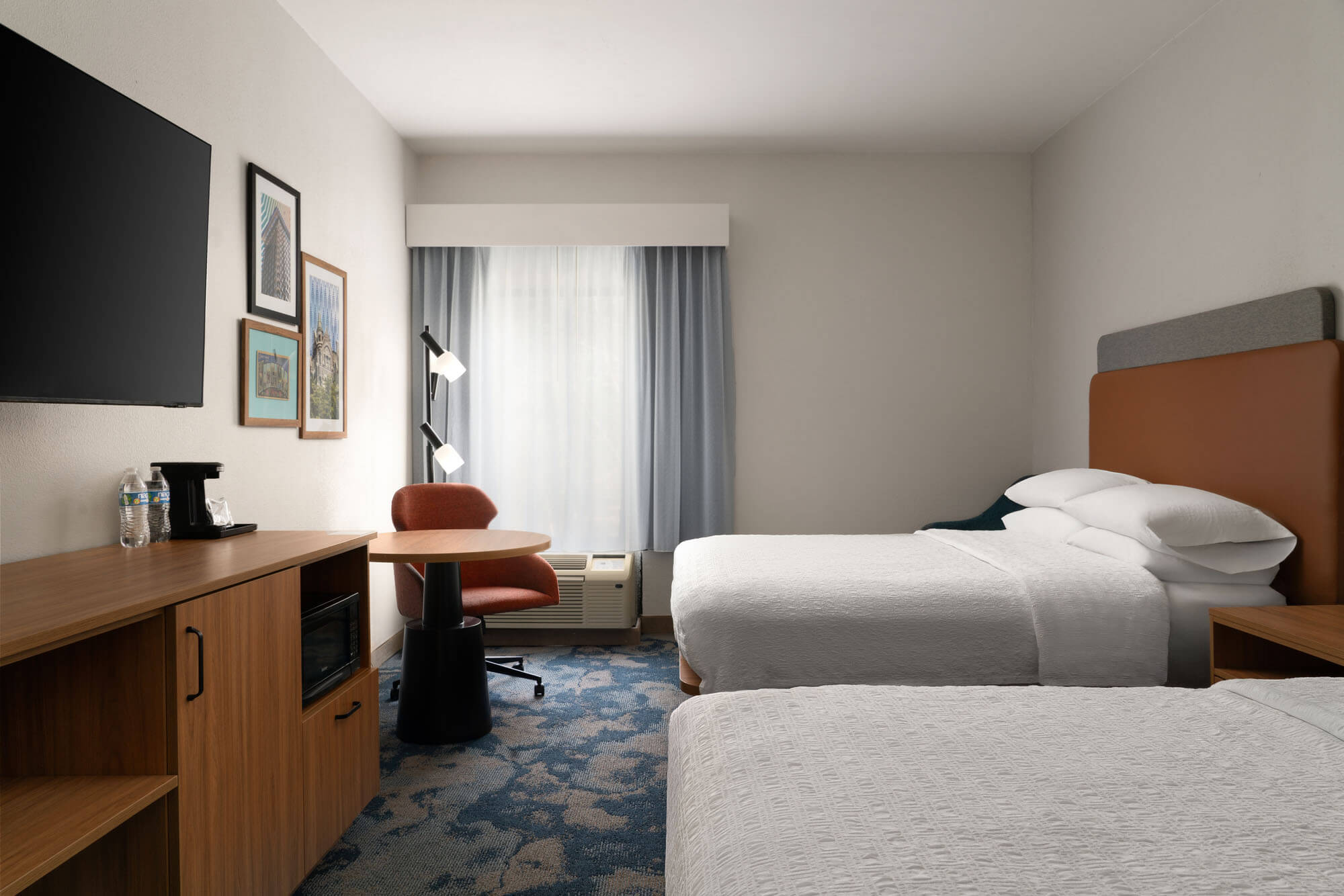 Hotel room with two twin beds, a wooden desk, red chair, wall art, and a window with blue curtains.