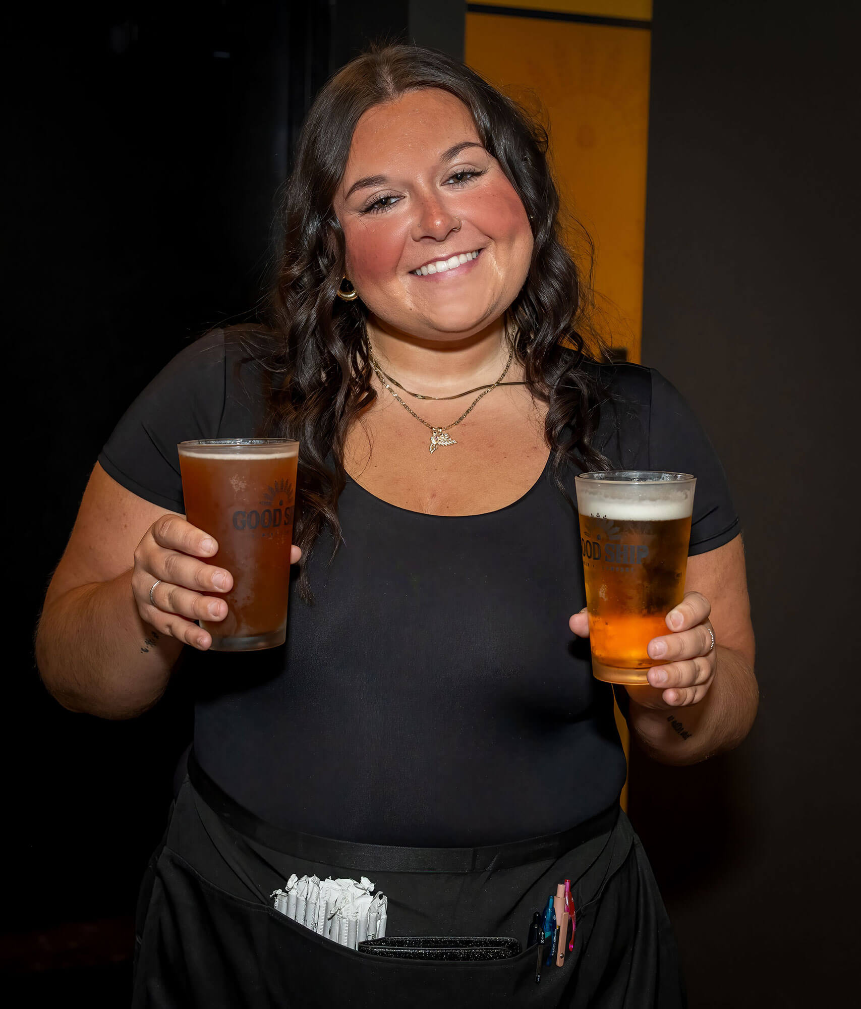 Good-Ship-Waitress → Visit Hopewell / Prince George VA Smiling woman in a black top holds a tall glass of beer in each hand, standing against a dark background.