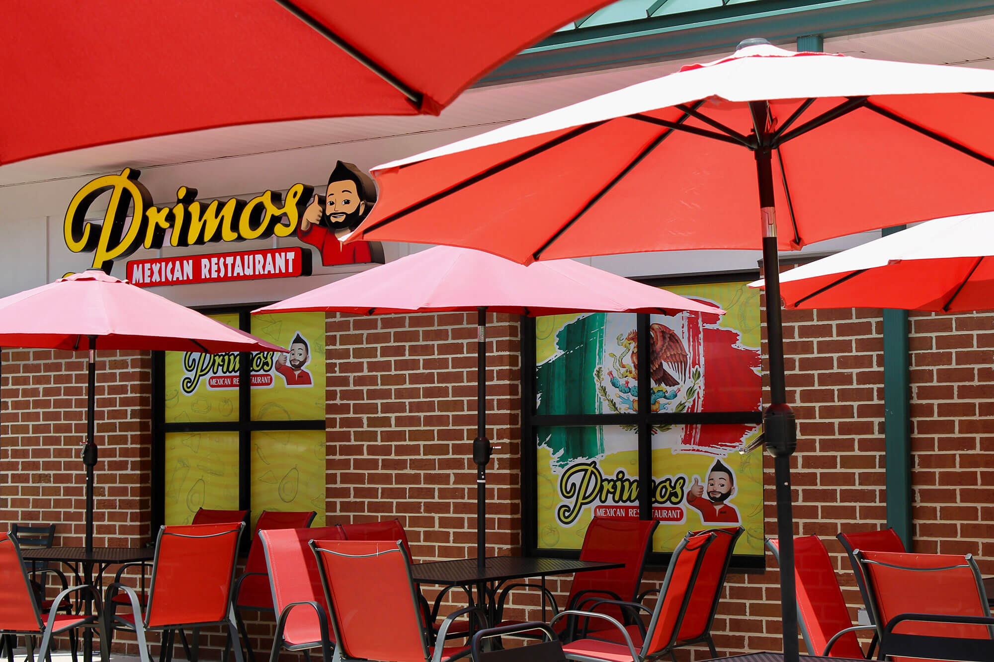 Outdoor seating area at Primos Mexican Restaurant with red patio umbrellas and red chairs against a brick wall exterior.