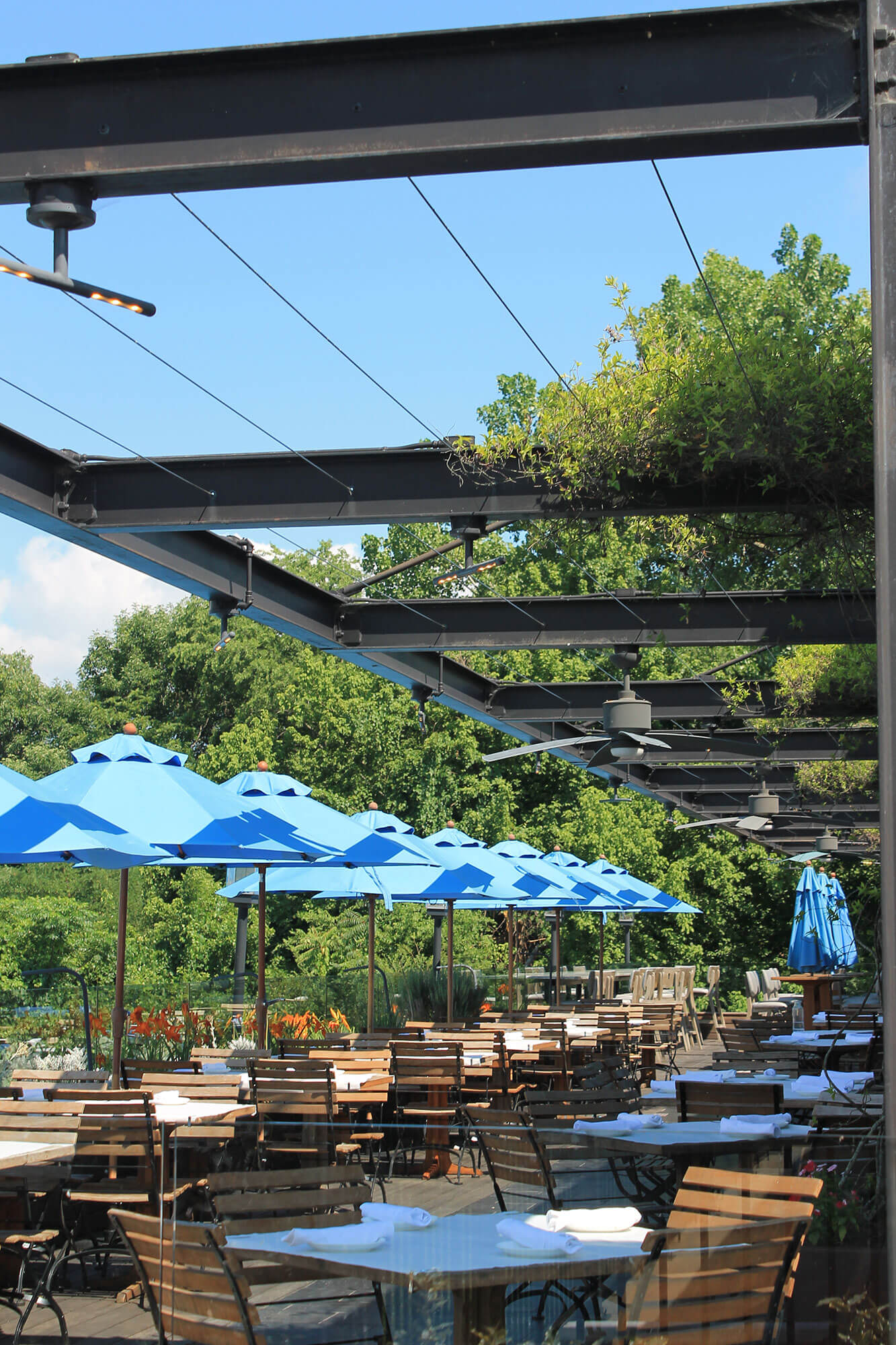 IMG_7683 → Visit Hopewell / Prince George VA → Visit Hopewell / Prince George VA Outdoor restaurant patio with wooden tables and chairs under bright blue umbrellas and a steel overhead structure.