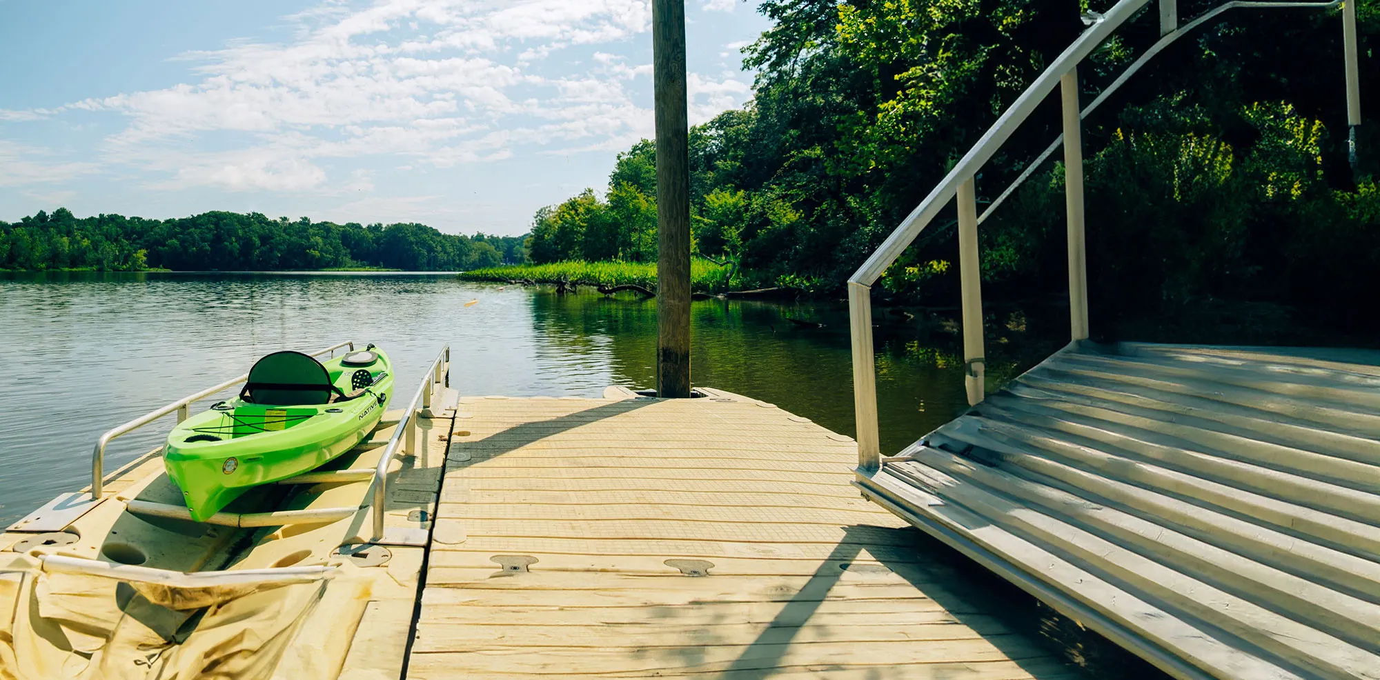 River-Kayak-wide → Visit Hopewell / Prince George VA → Visit Hopewell / Prince George VA Bright green kayak on a floating dock by a calm lake, with a wooden pier and stair railing on the right and green trees in the background.