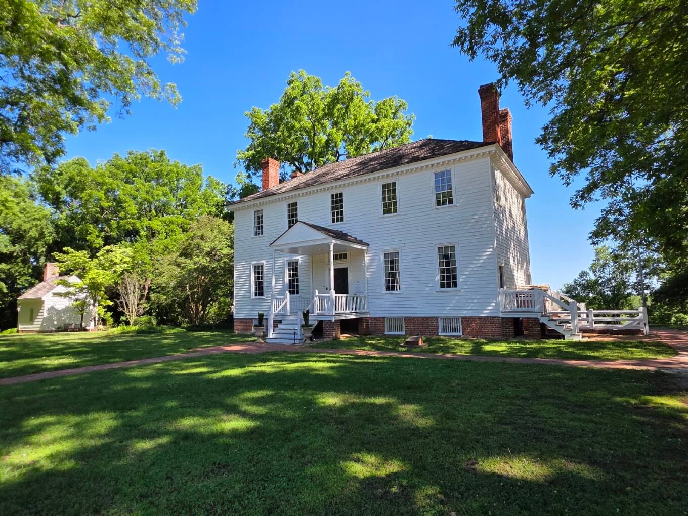 Front view of a white two-story colonial house with a small porch, brick foundation, and green lawn under a bright blue sky.