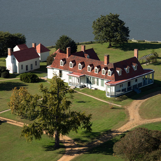 appomattox_manor → Visit Hopewell / Prince George VA → Visit Hopewell / Prince George VA Aerial view of a white, long red-roofed estate with multiple chimneys by a lake, surrounded by green lawns and a large tree in the foreground.