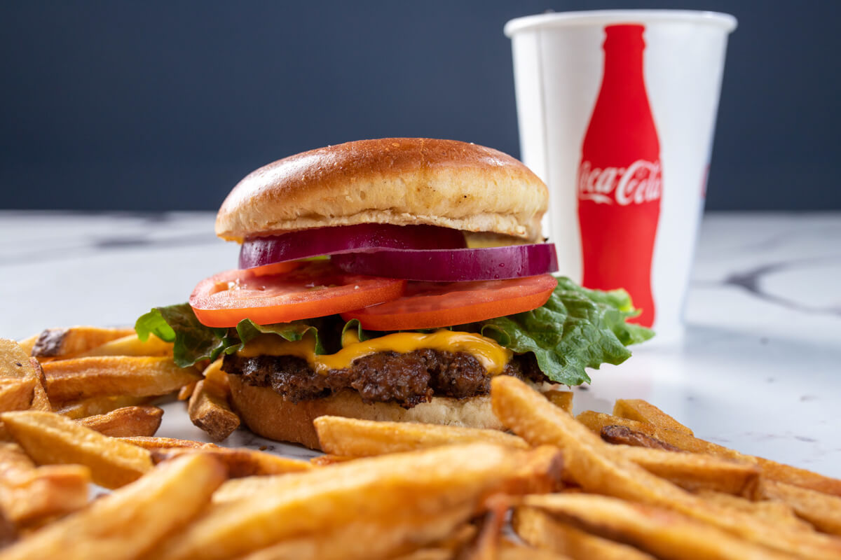 Juicy cheeseburger with lettuce, tomato, and red onion on a toasted bun, with fries on the side and a Coca-Cola cup in the background