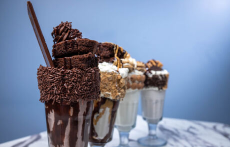 city-point-ice-cream-burgers → Visit Hopewell / Prince George VA → Visit Hopewell / Prince George VA Row of chocolate milkshakes topped with whipped cream and chocolate sprinkles, each with a tall chocolate stick against a blue background.