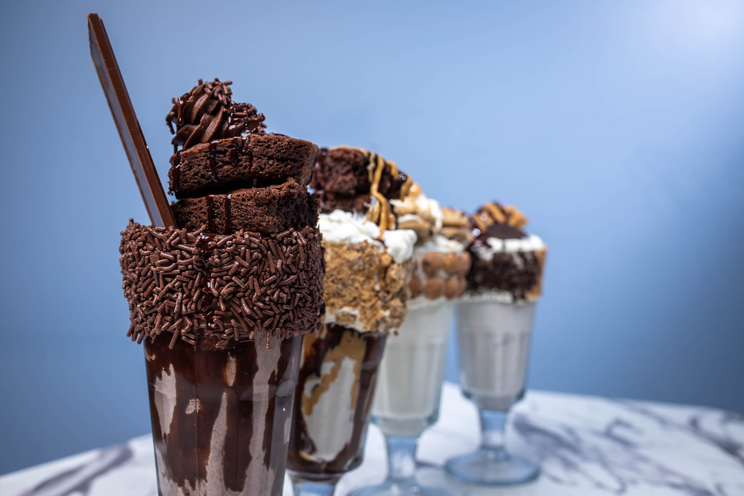 city-point-ice-cream-burgers → Visit Hopewell / Prince George VA → Visit Hopewell / Prince George VA Row of chocolate milkshakes topped with whipped cream and chocolate sprinkles, each with a tall chocolate stick against a blue background.