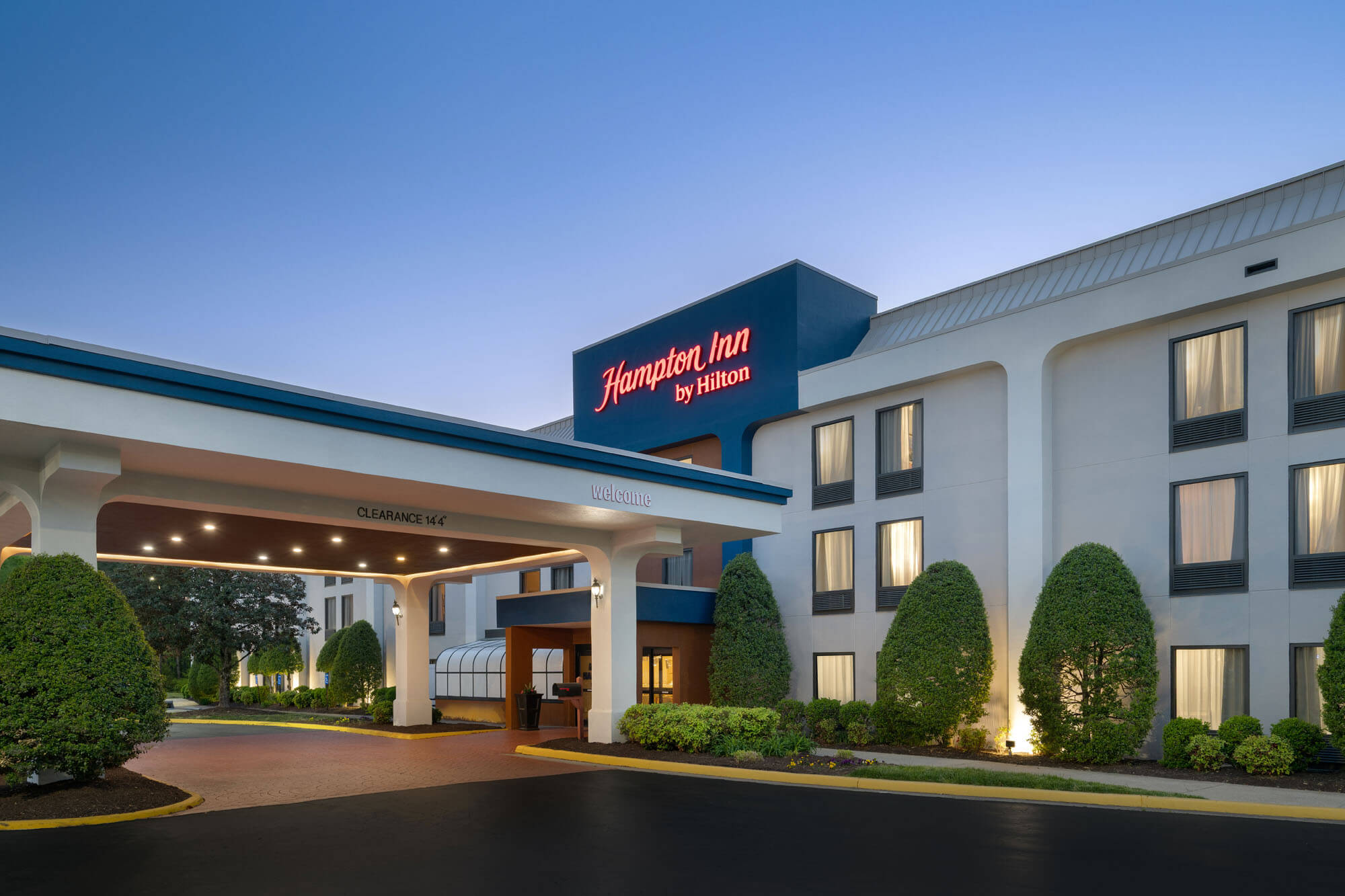 Exterior view of a Hampton Inn by Hilton hotel with a blue sign over the entrance and manicured shrubs along the front.