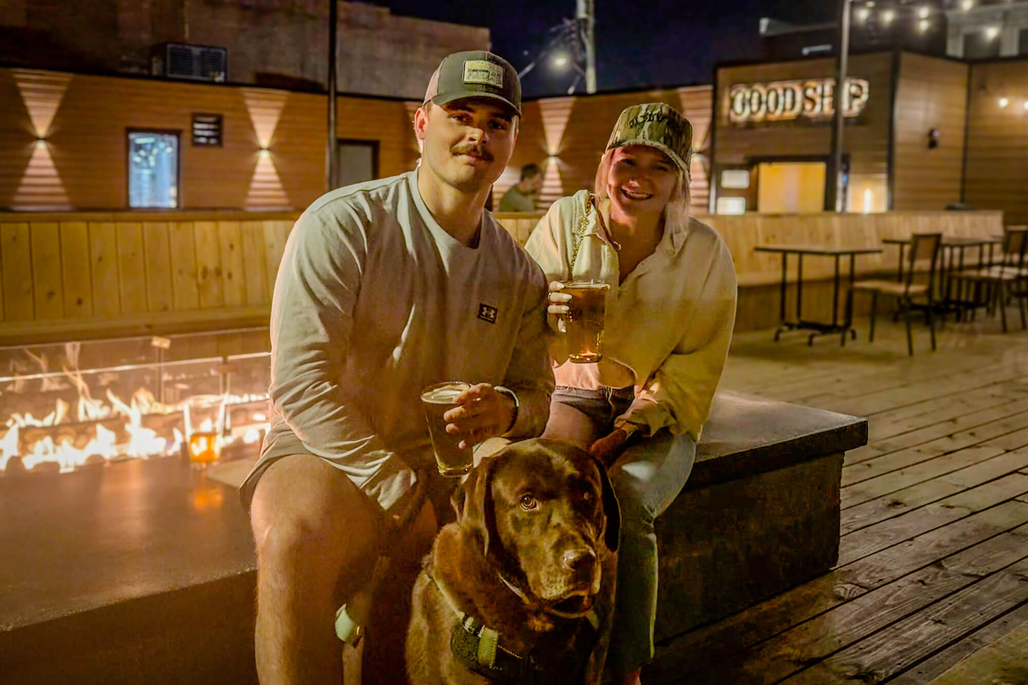 good-ship-couple-dog-beers → Visit Hopewell / Prince George VA Two friends sit by a glowing outdoor fire pit at night, holding beer glasses and smiling with a dog in front of them on a wooden deck.
