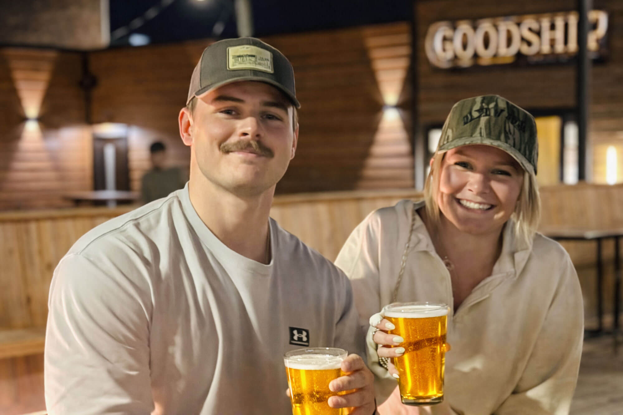 good-ship-happy → Visit Hopewell / Prince George VA → Visit Hopewell / Prince George VA Two friends smiling and toasting with beers at a wooden-bar pub setting.