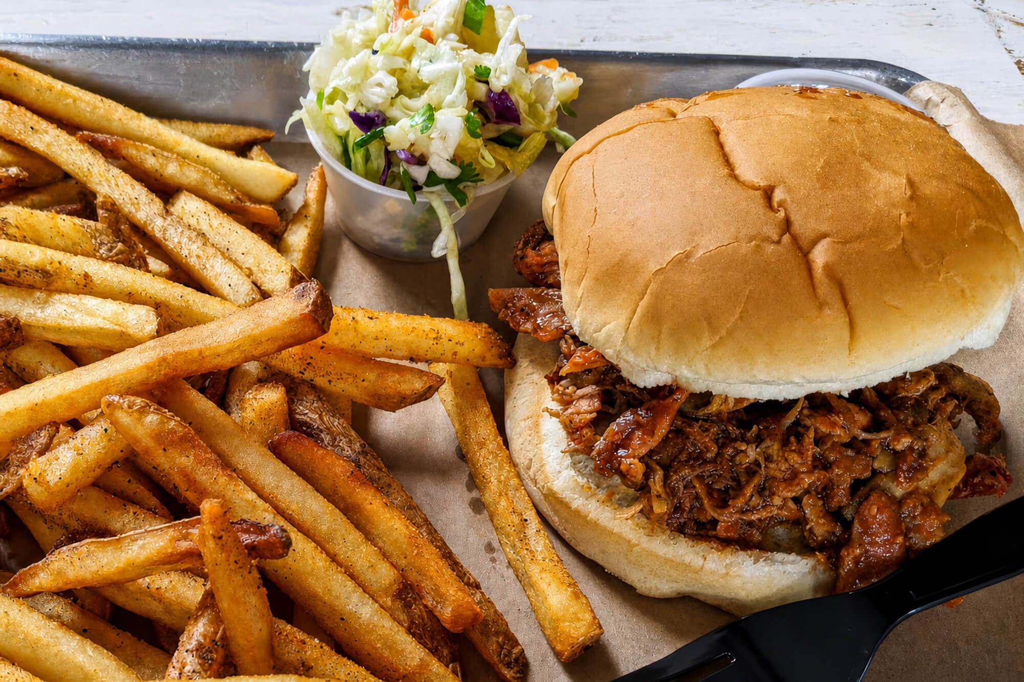 Hamburger with a large bun and shredded meat, surrounded by seasoned French fries and a small mixed-salad cup on a tray.