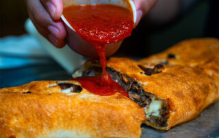 specials_back → Visit Hopewell / Prince George VA → Visit Hopewell / Prince George VA Person pours red sauce from a cup onto a golden-brown stuffed pastry with melted cheese and meat filling.