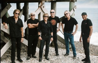 Beaver Brown → Visit Hopewell / Prince George VA Seven-member rock band posing on a pebbled beach under a wooden pier, all dressed in black.