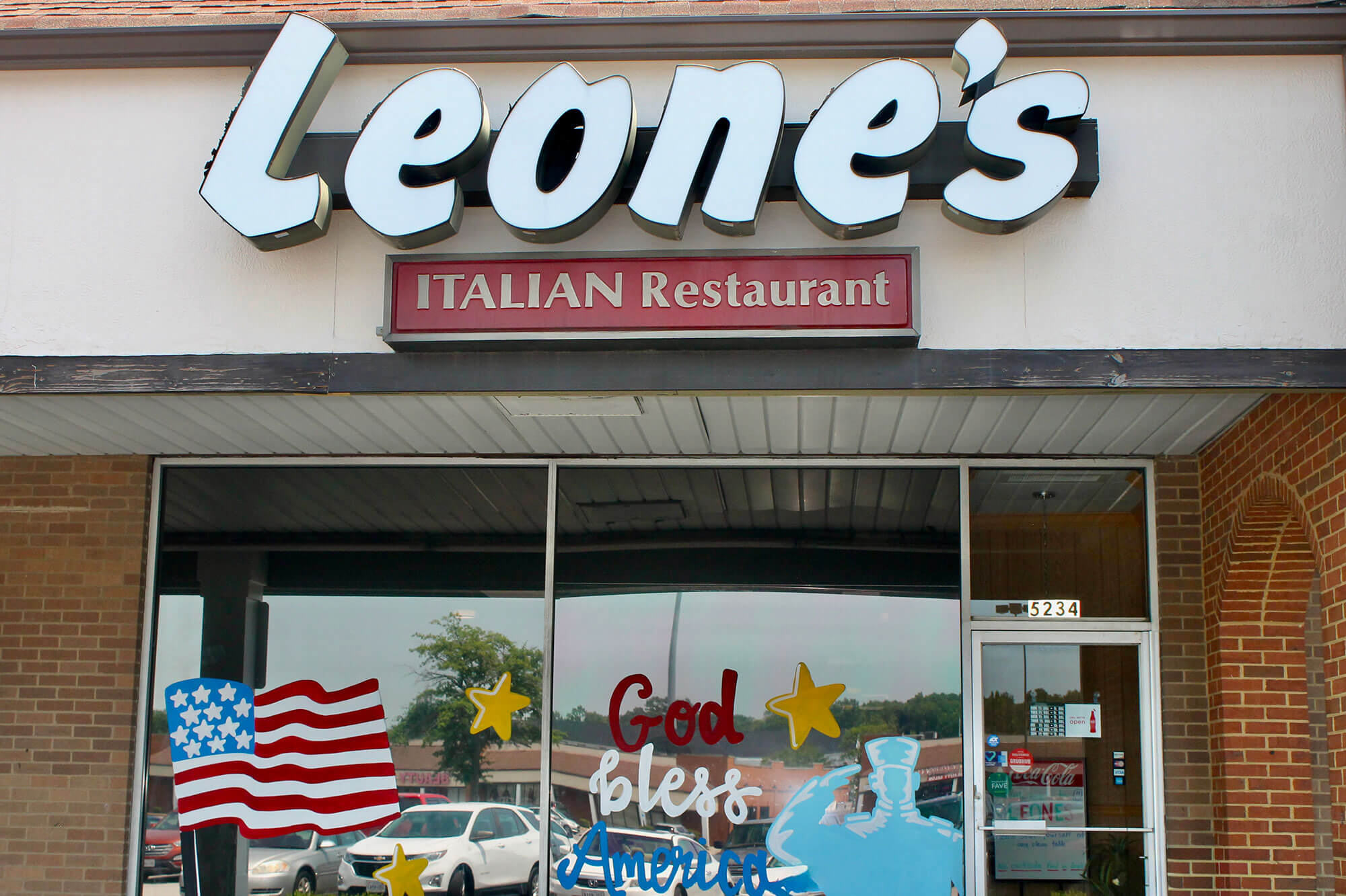 Storefront for Leone's Italian Restaurant with a red sign reading ITALIAN Restaurant above the door, brick facade, and patriotic window artwork showing an American flag and 'God Bless America' text.